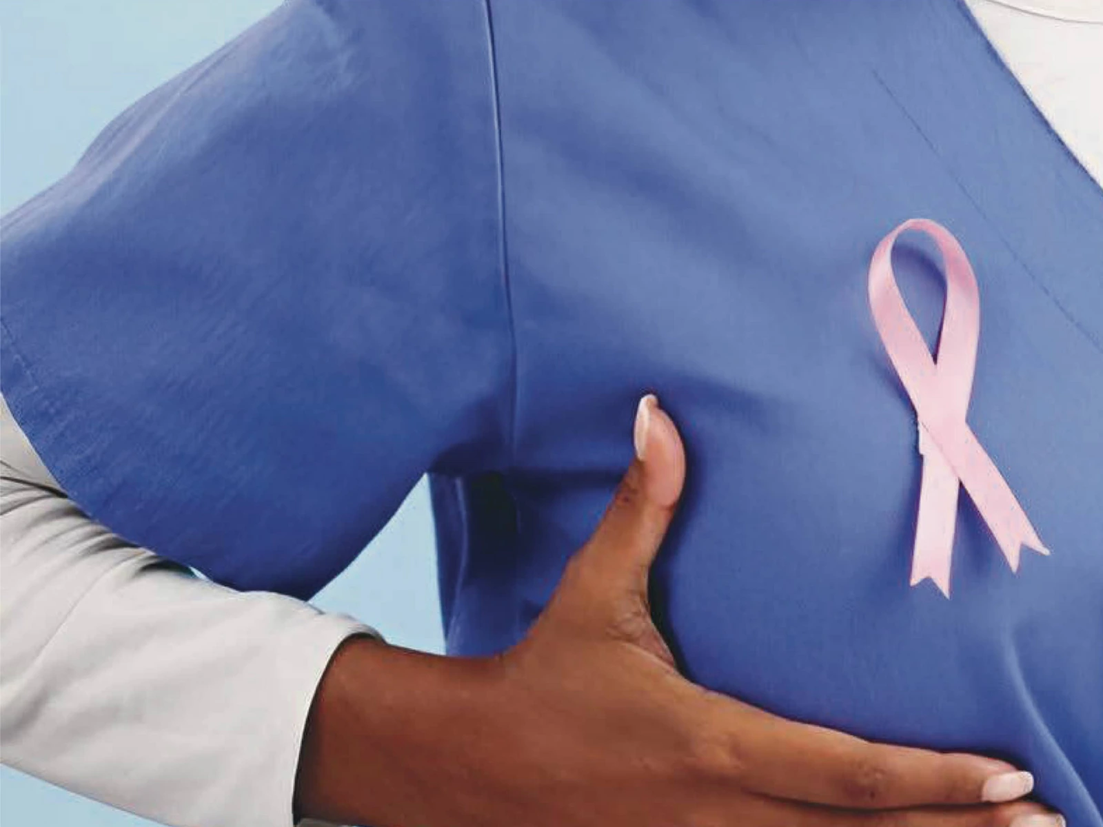 A nurse in blue scrubs wearing a pink breast cancer awareness ribbon, with a hand resting on the chest